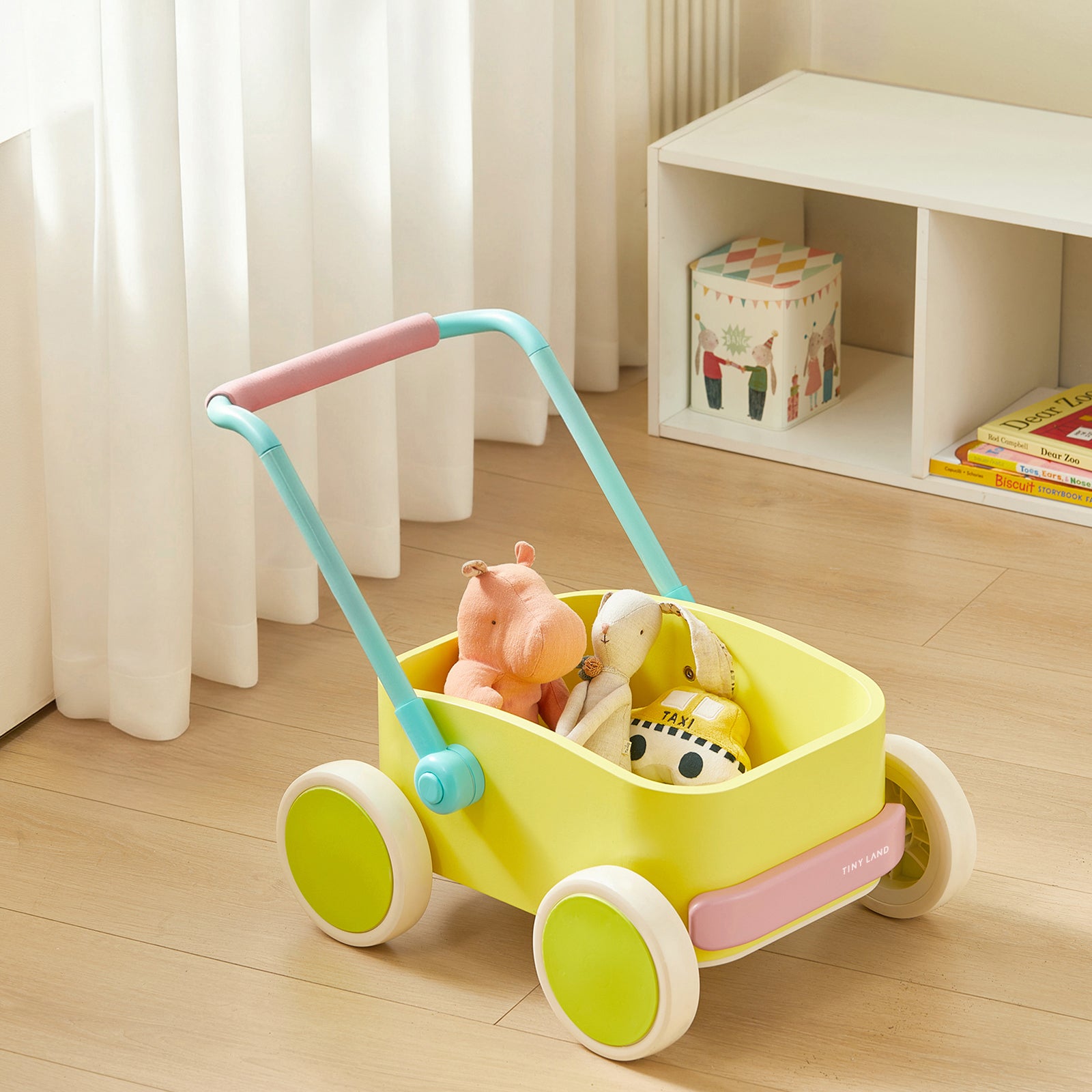 The Tiny Land® 3-in-1 Wooden Baby Walker Wagon in spring colors holds three plush toys—a hippo, a dog, and a giraffe—on a wooden floor near a white shelf with colorful children's books, toys, and a storage box.