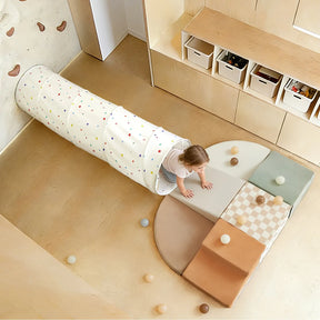A young child crawls out of the Tiny Land® Adventure Play Tunnel - Colorful Polka Dots onto soft play mats, with scattered balls in a modern playroom featuring wooden shelves and storage boxes.