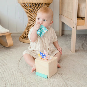 A baby in a light knit outfit sits on a carpeted floor, pulling colorful fabric squares from the Tiny Land® Montessori Toys for Newborns (4–6 Months) set, enhancing fine motor skills, with wicker furniture in the background.