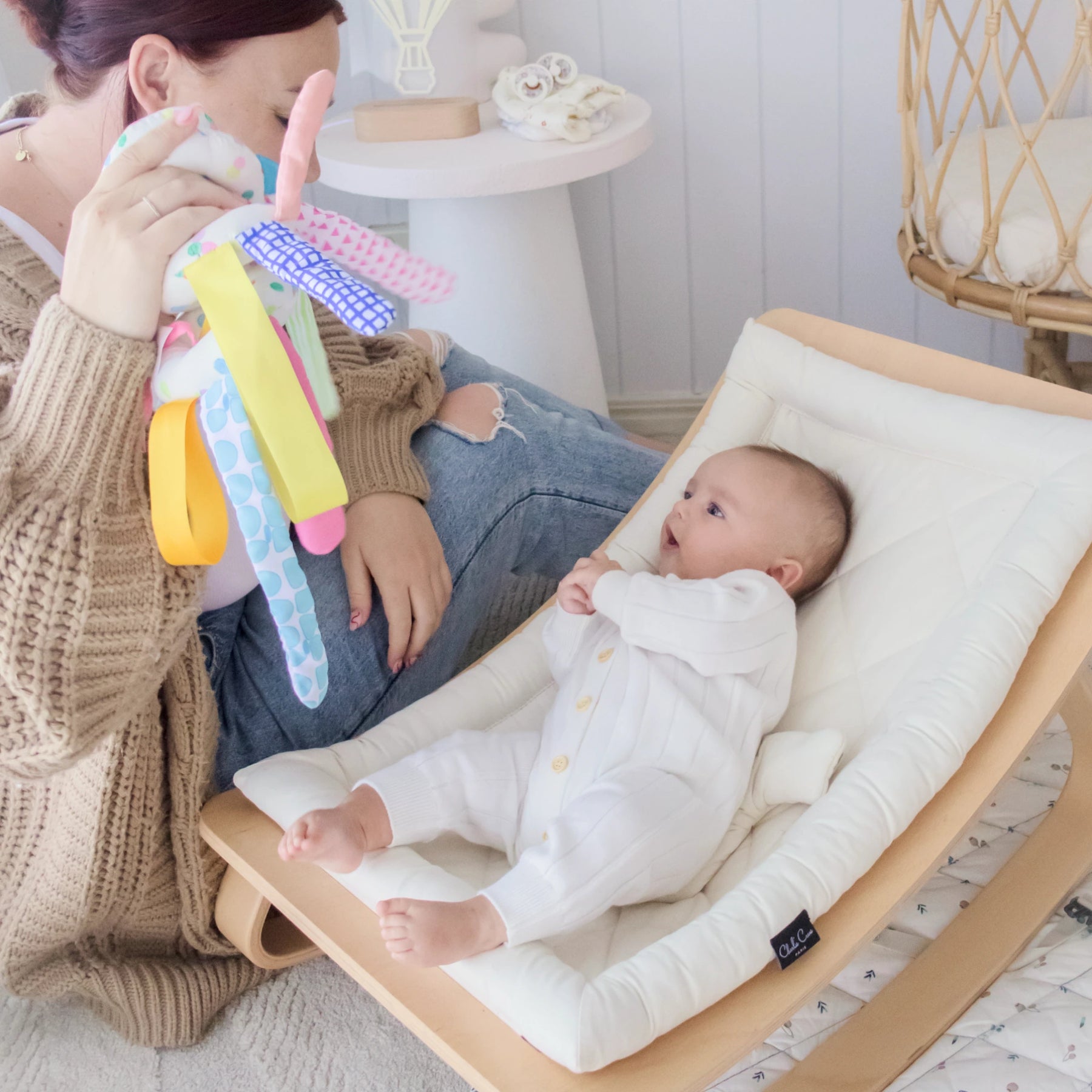 A woman sits on the floor, holding up a colorful Tiny Land® Montessori Toy for Newborns (0–3 Months) as her baby lies in a cream-colored rocker, watching with curiosity and enjoying sensory play in a cozy, well-lit room.