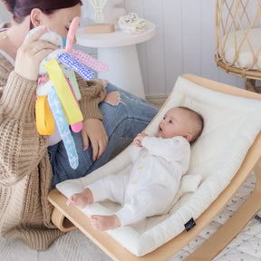 A woman sits on the floor, holding up a colorful Tiny Land® Montessori Toy for Newborns (0–3 Months) as her baby lies in a cream-colored rocker, watching with curiosity and enjoying sensory play in a cozy, well-lit room.