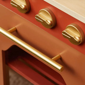 Close-up of the Tiny Land® Mediterranean Play Kitchen by Tiny Land, featuring a red and gold oven with three gold knobs, a gold oven handle, and a light wood surface above|Mediterranean