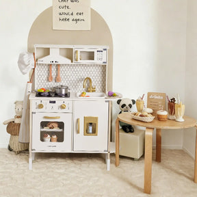 The Tiny Land® PopJoy Play Kitchen by Tiny Land sits against a beige wall, with pots, pans, and utensils. A small round table with toy food and a panda plush is nearby, while stuffed animals surround the play kitchen for pretend fun.