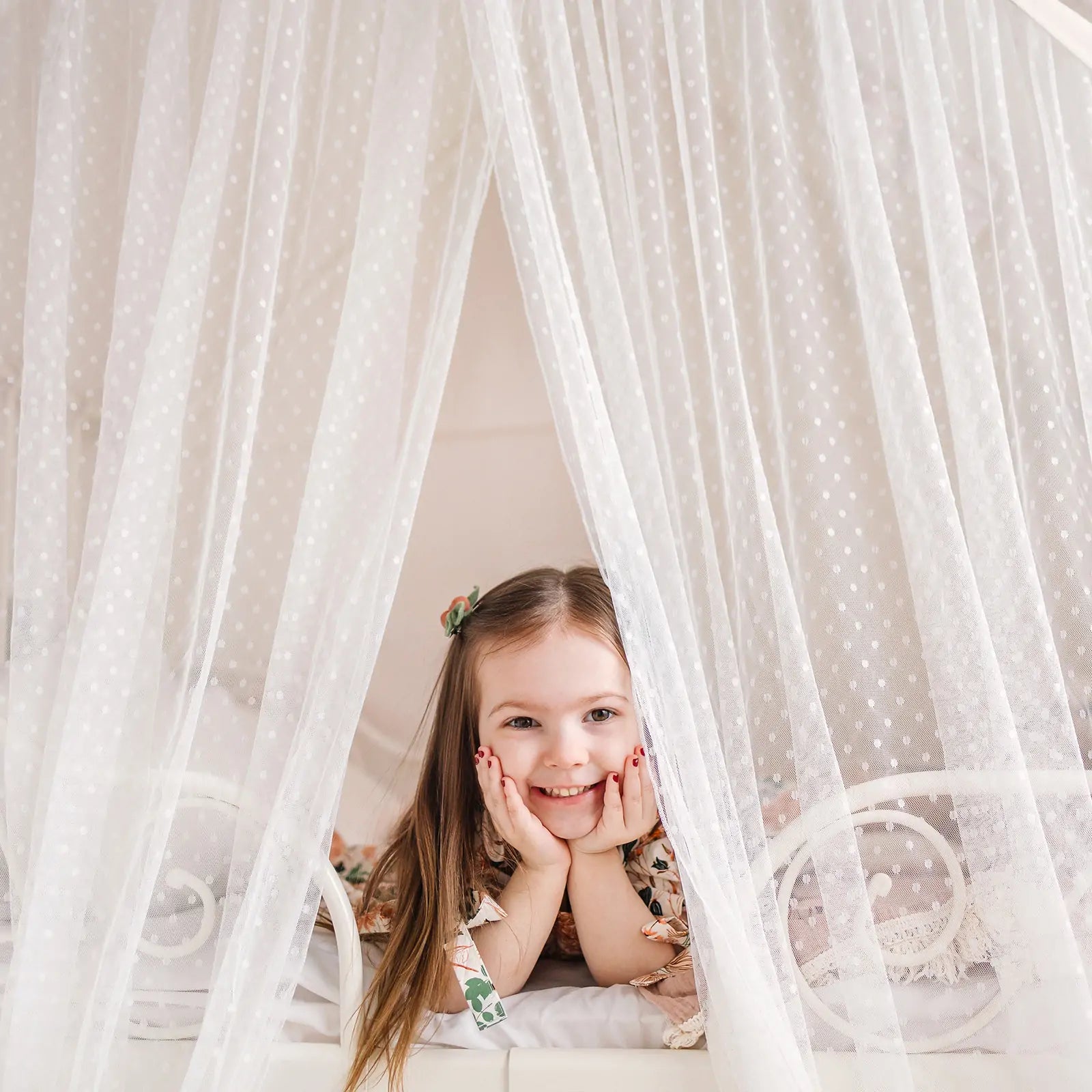 A young girl with long brown hair smiles as she rests on a bed inside the Tiny Land® Large Space Play House with Star Lights, surrounded by sheer white dotted curtains for a cozy, tent-like feel.|mesh