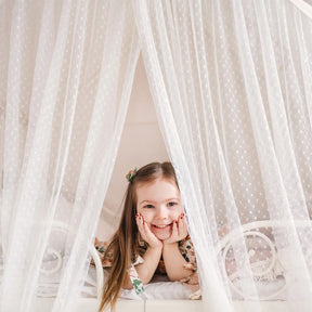 A young girl with long brown hair smiles as she rests on a bed inside the Tiny Land® Large Space Play House with Star Lights, surrounded by sheer white dotted curtains for a cozy, tent-like feel.|mesh