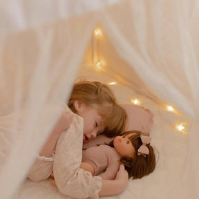A young child relaxes under the Tiny Land Bright Play & Art Set—featuring a colorful easel and a cream playhouse with lights—enjoying the warm glow and cozy, dreamy atmosphere created by soft fairy lights inside the tent|white