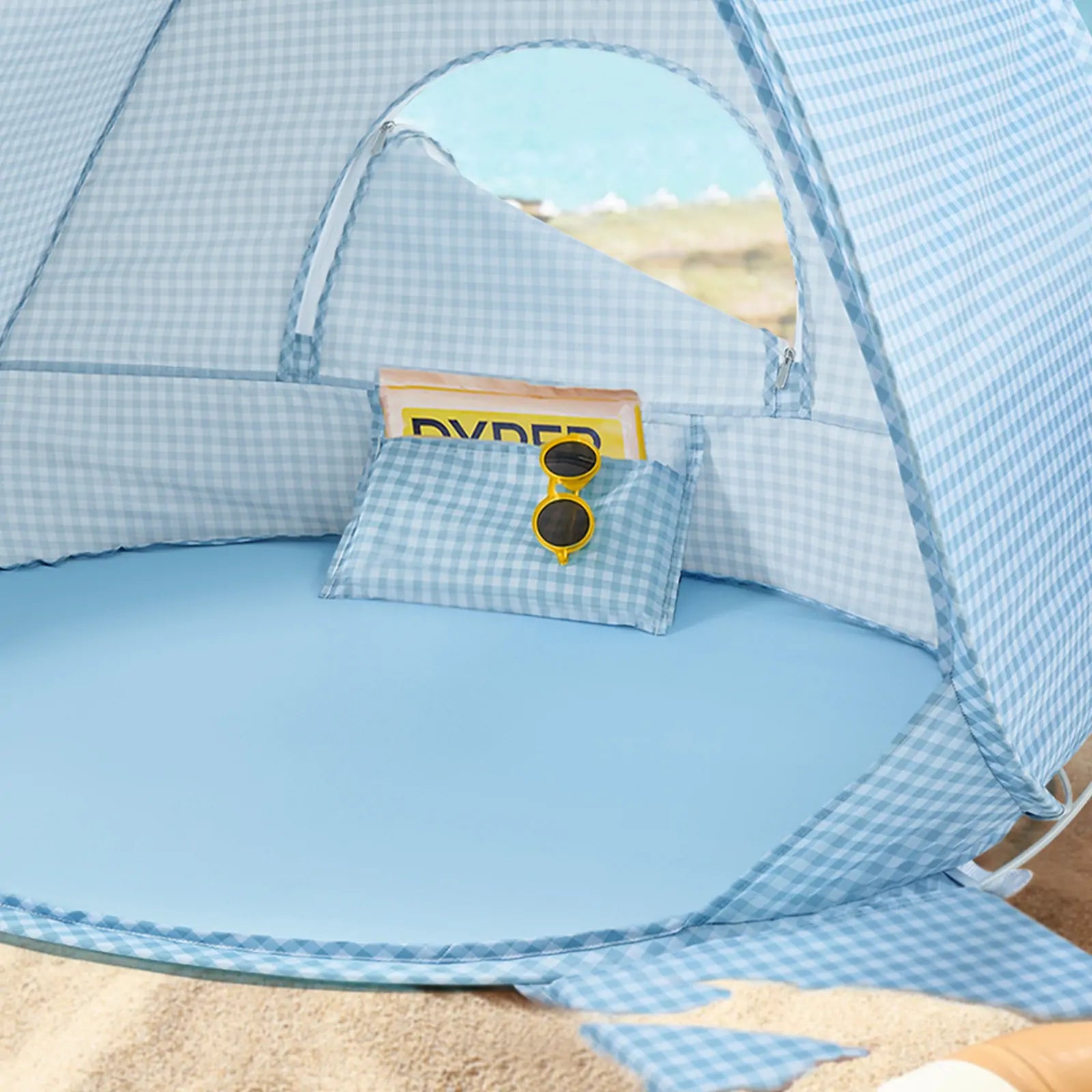 The Tiny Land® One-Stop Beach Toy & Tent Set in light blue gingham sits on the sand, complete with a matching pillow, yellow sunglasses, and magazine. Relax under this UV-protective tent and enjoy your sea view in comfort.|Light Blue Gingham
