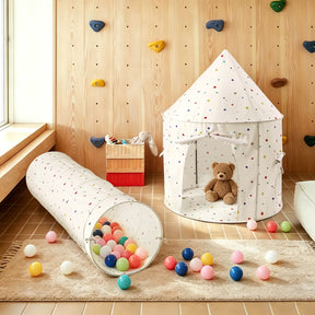A cozy playroom with a wooden wall and climbing holds, a Tiny Land® Adventure Play Tunnel - Colorful Polka Dots, colorful balls on the floor, a teddy bear in the tunnel, and a toy basket in the background.