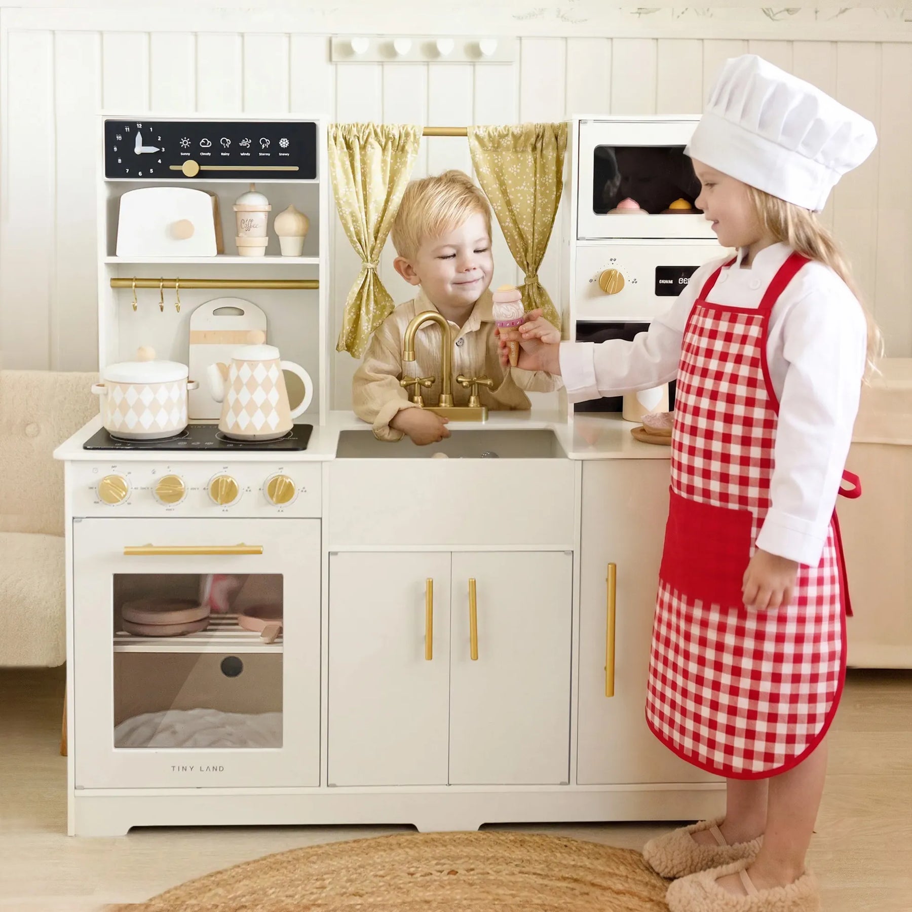 Two children play with the Tiny Land® Iconic Wooden Play Kitchen – Cream Pretend Cooking Set for Kids. One, in a chef’s hat and apron, serves food while the other smiles behind the pretend sink. The Tiny Land kitchen includes appliances and fun accessories.