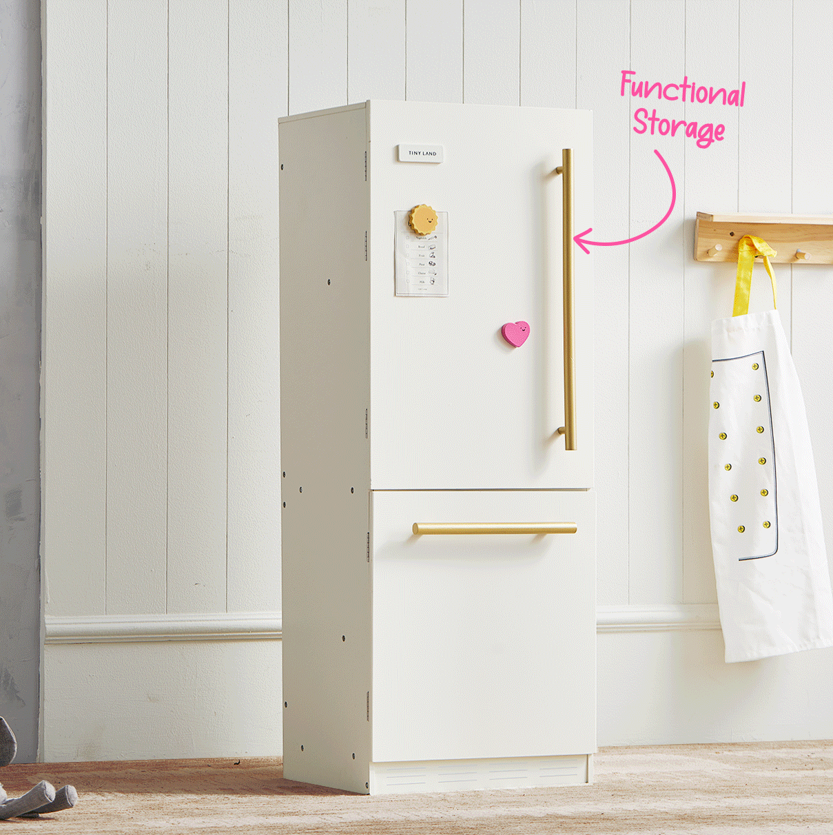 A white Tiny Land® Iconic Water Play Kitchen & Premium Wooden Refrigerator by Tiny Land INC stands against a white paneled wall, resembling a charming wooden cabinet perfect for pretend play. A pink heart magnet and paper are on the door, while an apron hangs on a wooden rack nearby. The words “Functional Storage” with an arrow point toward the refrigerator.