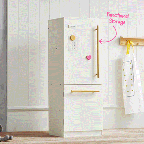 A white Tiny Land® Iconic Water Play Kitchen & Premium Wooden Refrigerator by Tiny Land INC stands against a white paneled wall, resembling a charming wooden cabinet perfect for pretend play. A pink heart magnet and paper are on the door, while an apron hangs on a wooden rack nearby. The words “Functional Storage” with an arrow point toward the refrigerator.