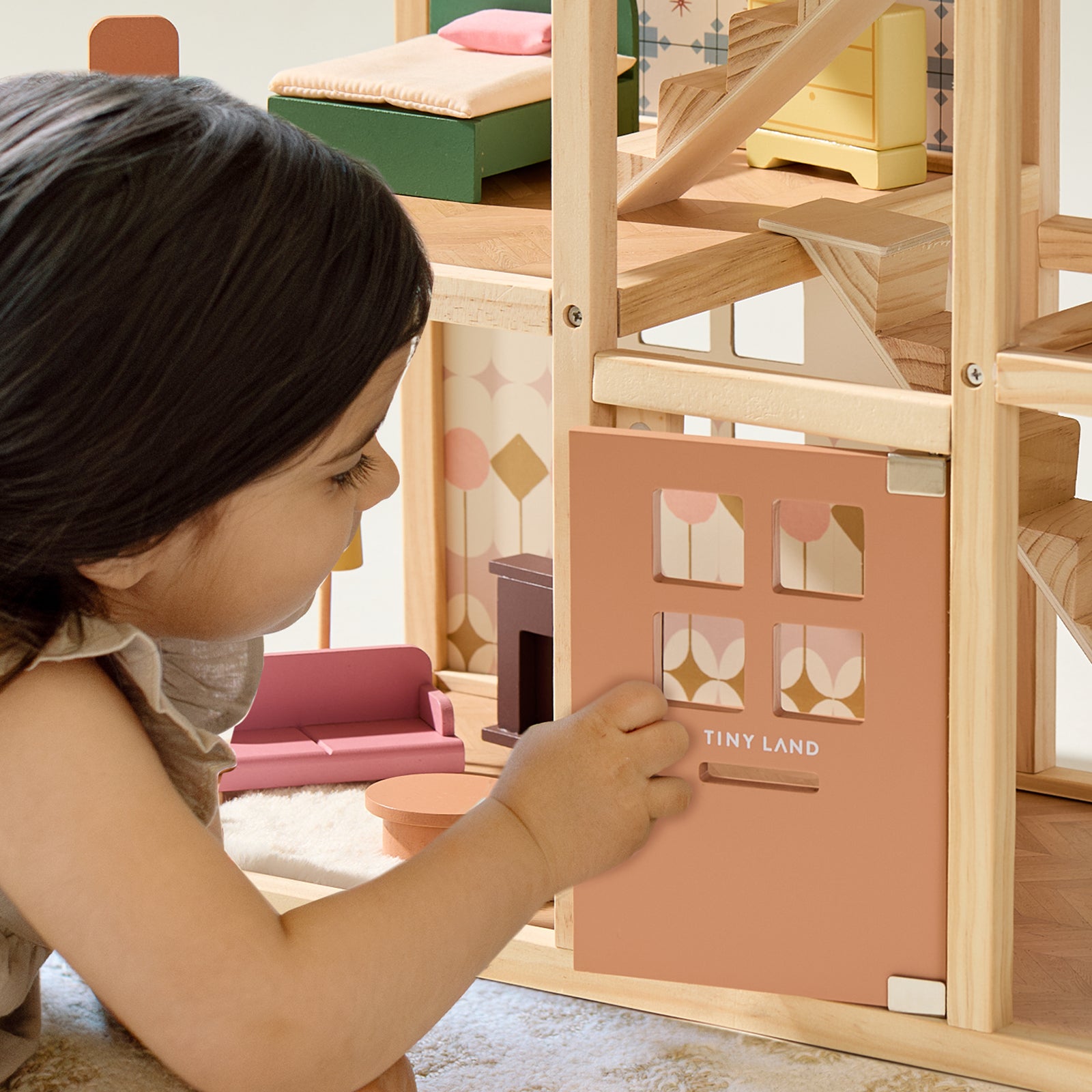A young child enjoys imaginative play with the Tiny Land® Sweetwood Dollhouse without Dolls, complete with miniature furniture sets and patterned wallpaper behind its peach-colored door labeled Tiny Land.|Pegasus