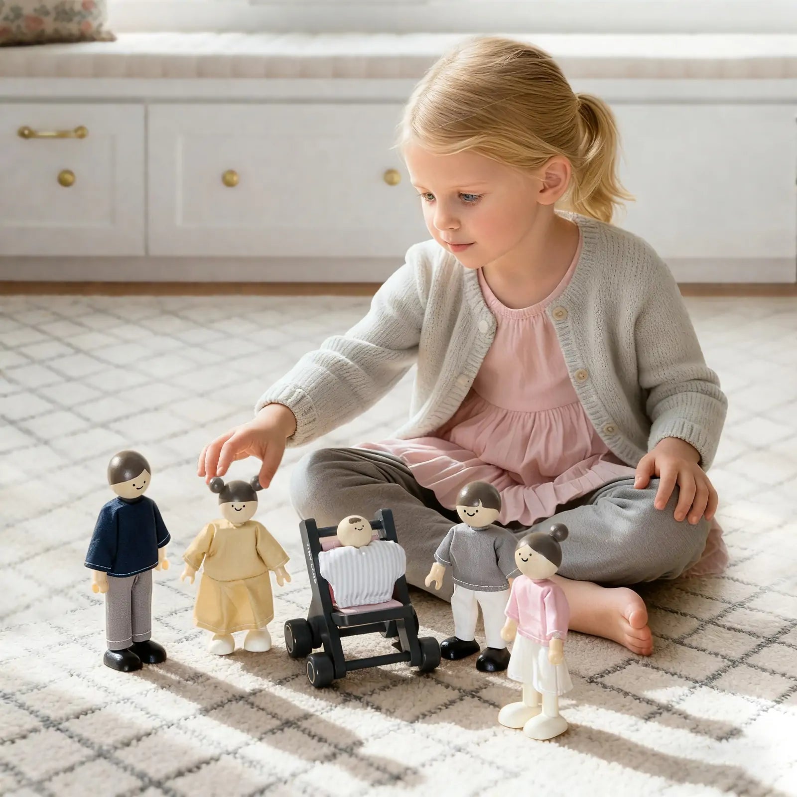 A young girl in a light pink dress and gray cardigan plays on a carpeted floor with Tiny Land® Wooden Dollhouse Dolls, including a baby doll in a stroller, filling the bright, cozy room with charm.