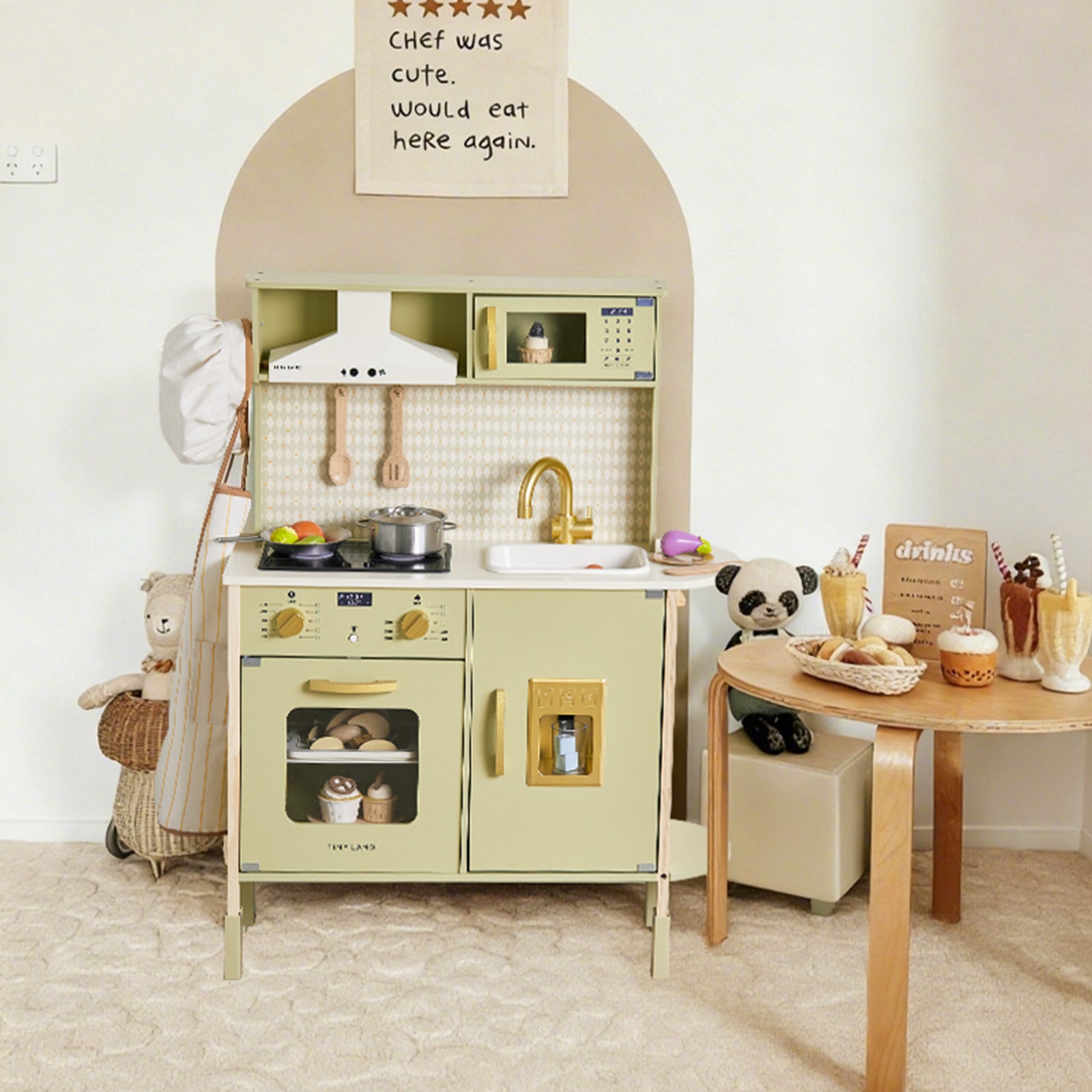 A delightful scene features the Tiny Land® PopJoy Play Kitchen in Pink / Green by Tiny Land, a wooden toy kitchen set adorned with play pots, pans, and pretend food. Beside it stands a small table displaying make-believe drinks and desserts, while plush stuffed animals assemble for lively pretend play. A whimsical sign on the wall proclaims: THE CHEF WAS CUTE. would eat here again.|Sage