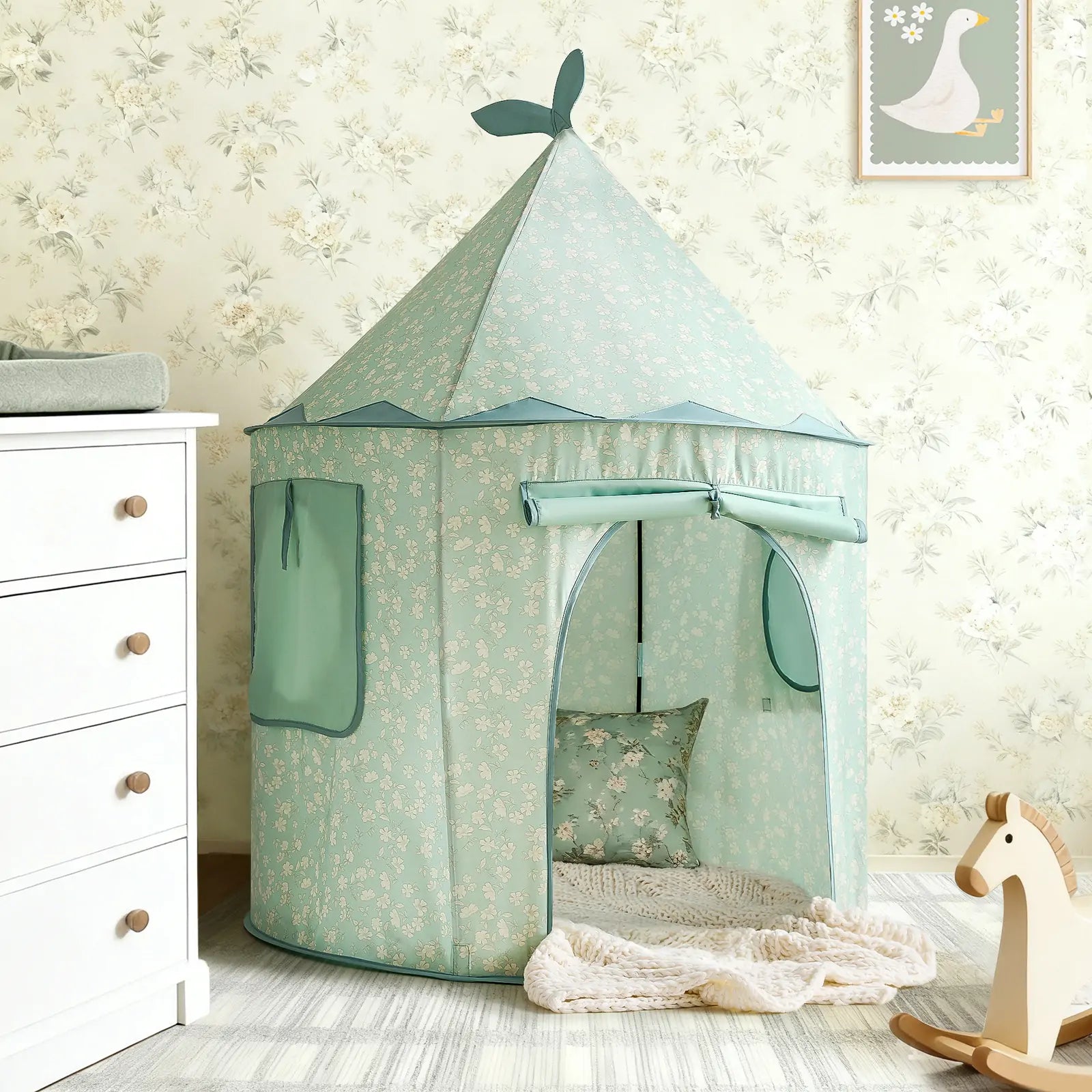 The Tiny Land® Tiny Sprout Castle Tent in mint green, featuring a pointed roof and leaf accents, stands in a child’s room with floral wallpaper, near a wooden rocking horse, white dresser, and framed goose picture on the wall.|Green