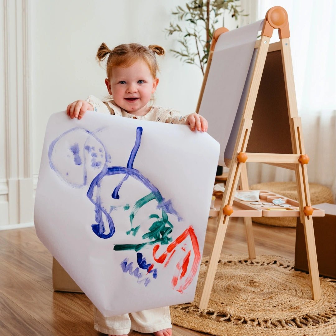 A young child with pigtails smiles while showing her colorful art, created on the Tiny Land® Caramel Magnitales Easel for Kids. The sunlit room and assorted supplies set the scene for creative fun with this Tiny Land essential.