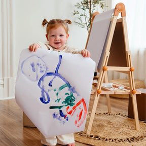 A young child with pigtails smiles while showing her colorful art, created on the Tiny Land® Caramel Magnitales Easel for Kids. The sunlit room and assorted supplies set the scene for creative fun with this Tiny Land essential.