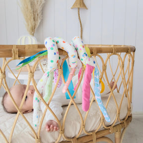 A baby lies in a wicker crib, reaching for colorful toys from the Tiny Land® Montessori Toys for Newborns (0–3 Months) by Tiny Land, draped over the crib’s edge in a bright nursery—perfect for early sensory play.