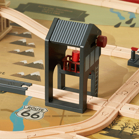 The Tiny Land® 1920 Railway Adventure Train Table features a wooden crossing signal with a black roof and red lever on tracks, set atop a Route 66 map-themed play mat with mountain graphics and 1920s city-inspired interactive elements.