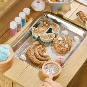 A child explores shaped sand, seashells, and molds from the Tiny Land® Outdoor Mud Kitchen & Sand Play Set on a wooden table, enhanced by glitter jars and decorative items from Tiny Land for endless outdoor play.