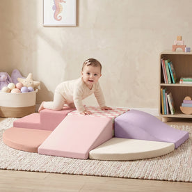 A baby in light clothing climbs on the pastel violet Tiny Land® PlayNest Climbing Blocks by Tiny Land in a cozy nursery, with a basket of toys, a small shelf, and wall art in the background.