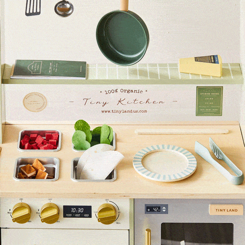 The Tiny Land® BistroSwitch Play Kitchen by Tiny Land includes a pan, utensils, faux veggies, plate, recipe cards, stovetop with dials and timer, and an accessory shelf. Great for pretend play! The text reads “100% Organic Tiny Kitchen.”.|Green