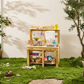 The Tiny Land® Outdoor Mud Kitchen & Sand Play Set by Tiny Land features a wooden kitchen on moss, with toy utensils, pots, cups, and play food on its shelves. A yellow watering can and toy tools from the set hang above the workspace near a leafy plant.