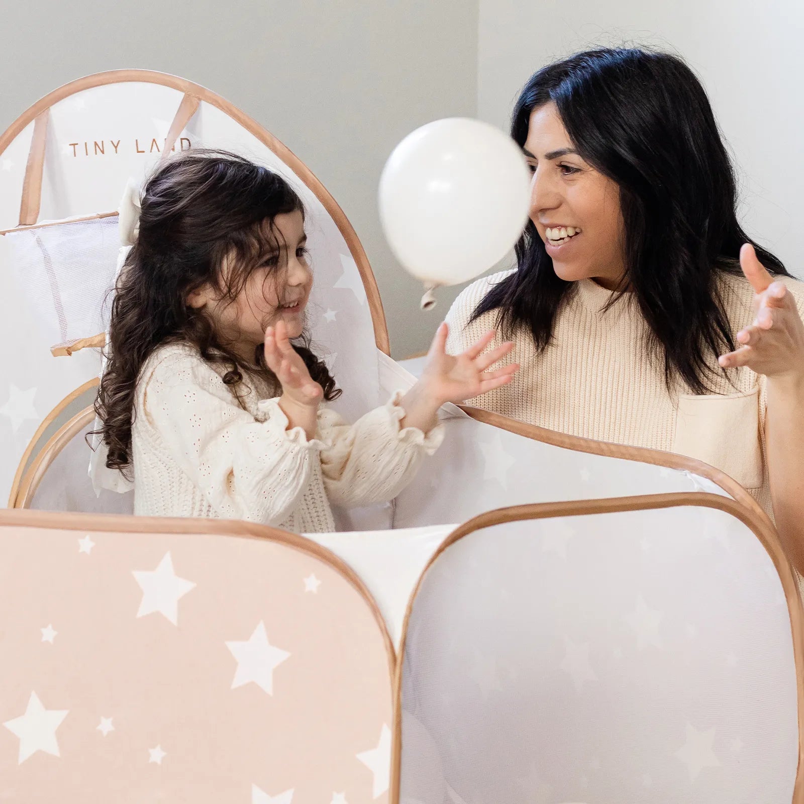 A smiling woman and young girl play with a white balloon inside the Tiny Land® Compact Adventure 3-in-1 Play Tunnel, their laughter filling the star-patterned tent with joy. Both look happy and engaged, sharing a delightful moment together as the nearby play tunnel from Tiny Land promises even more fun adventures.|brown