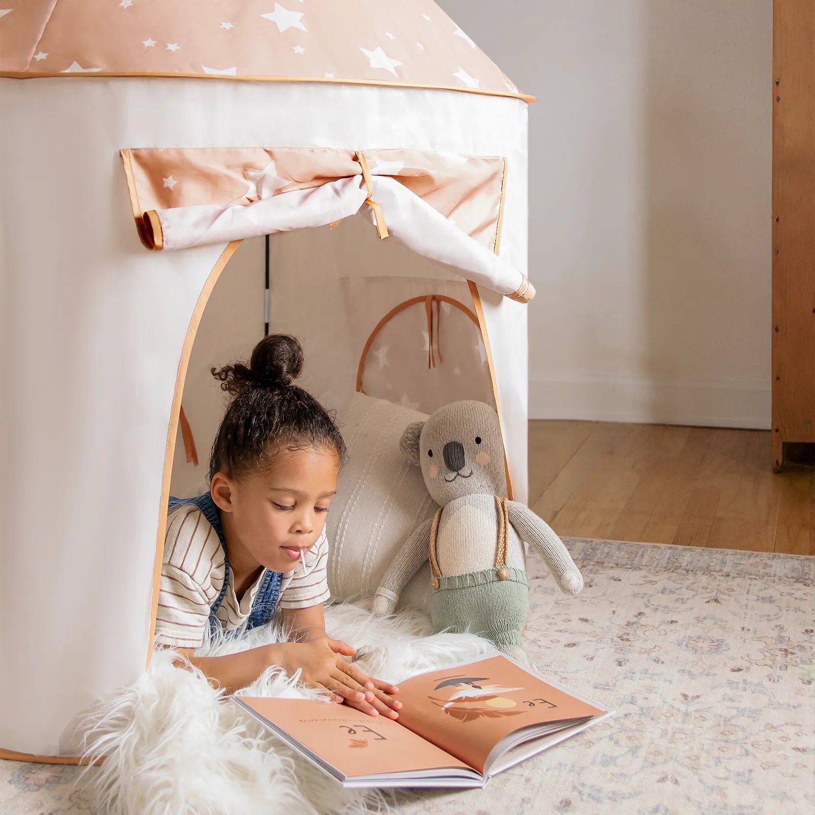 A young child reads a book while lying inside a star-patterned play tent, with a stuffed bear toy beside her on a soft rug in a cozy room. Nearby, the entrance to the Tiny Land® Compact Adventure 3-in-1 Play Tunnel adds to the playful atmosphere.|brown