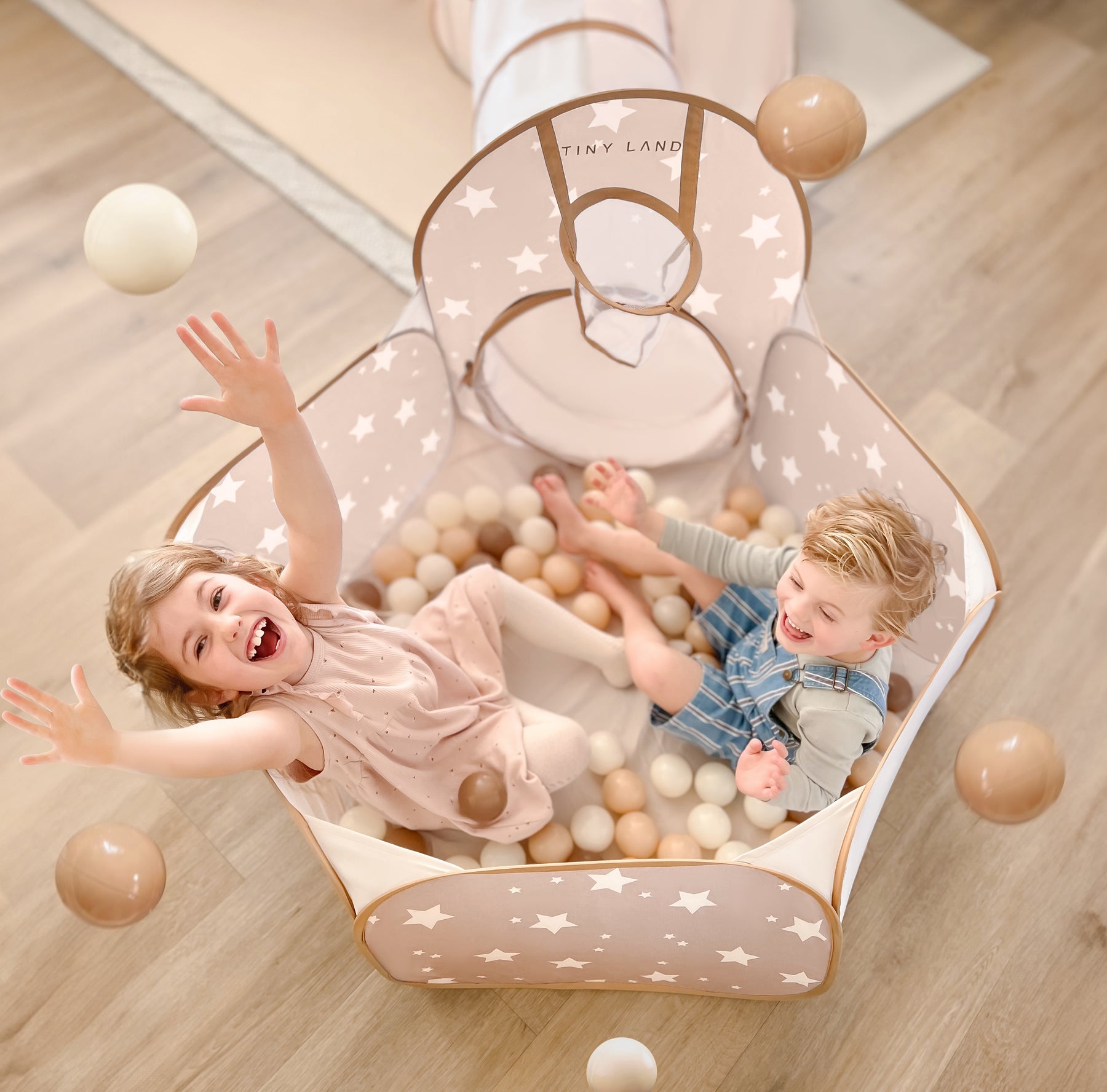 Two young children laugh and toss Tiny Land® Ocean Ball - 200 pcs with Varied Colors in a star-patterned, beige playpen filled with soft, non-toxic balls. A play tunnel attaches to the playpen on a light wood floor. Brand: Tiny Land.|Brown