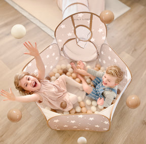 Two young children laugh and toss Tiny Land® Ocean Ball - 200 pcs with Varied Colors in a star-patterned, beige playpen filled with soft, non-toxic balls. A play tunnel attaches to the playpen on a light wood floor. Brand: Tiny Land.|Brown