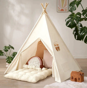 A Tiny Land® Thick Padded Play Cushion sits inside a cozy indoor teepee with pillows. Green plants, framed wall art, and a small woven house-shaped basket add charm to the inviting play area. Arch not included.