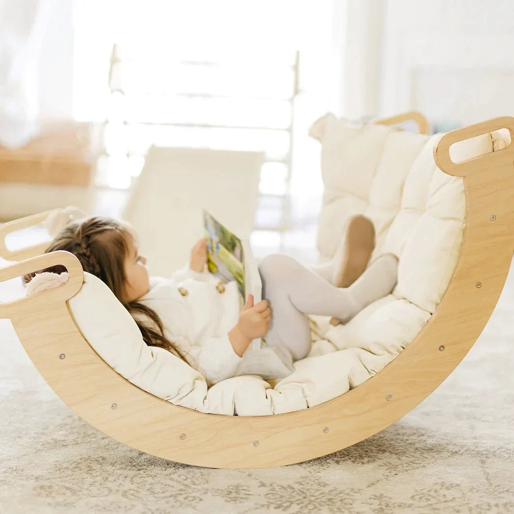 A young child reclines on a wooden rocker, reading, atop the Tiny Land® Thick Padded Play Cushion by Tiny Land. The scene is bright and cozy with soft natural light and a comfortable, light-colored setting.