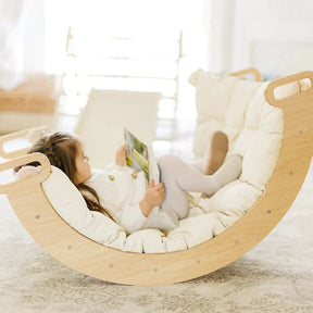 A young child reclines on a wooden rocker, reading, atop the Tiny Land® Thick Padded Play Cushion by Tiny Land. The scene is bright and cozy with soft natural light and a comfortable, light-colored setting.