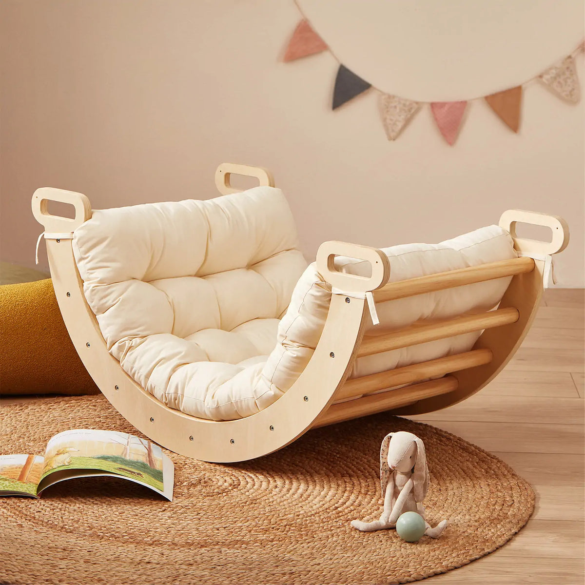 A Tiny Land® Thick Padded Play Cushion sits on a round woven rug beside a stuffed rabbit and an open children’s book, with colorful bunting in the background. The wooden rocking chair and Arch are not included.