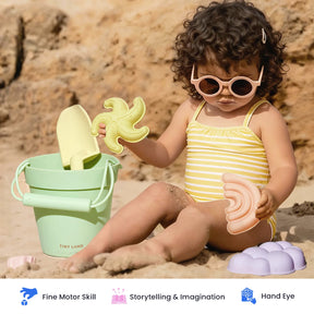 A young child in a yellow striped swimsuit and round sunglasses engages in sensory play with the Tiny Land® Sand-Buddies Sand Toy Set by Tiny Land—including a bucket, shovel, and pastel sand molds—while sitting on sandy ground near rocks.