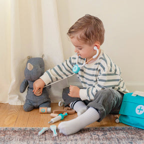 A young child in a striped shirt plays pretend doctor with the Tiny Land® Doctor Kit for Kids by Tiny Land, using a toy stethoscope to examine a stuffed animal on the floor.