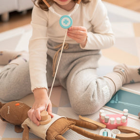 A child in comfy clothes pretends to check a plush toy with a stethoscope, surrounded by play tools from the Tiny Land® Doctor Kit for Kids by Tiny Land on a soft mat.
