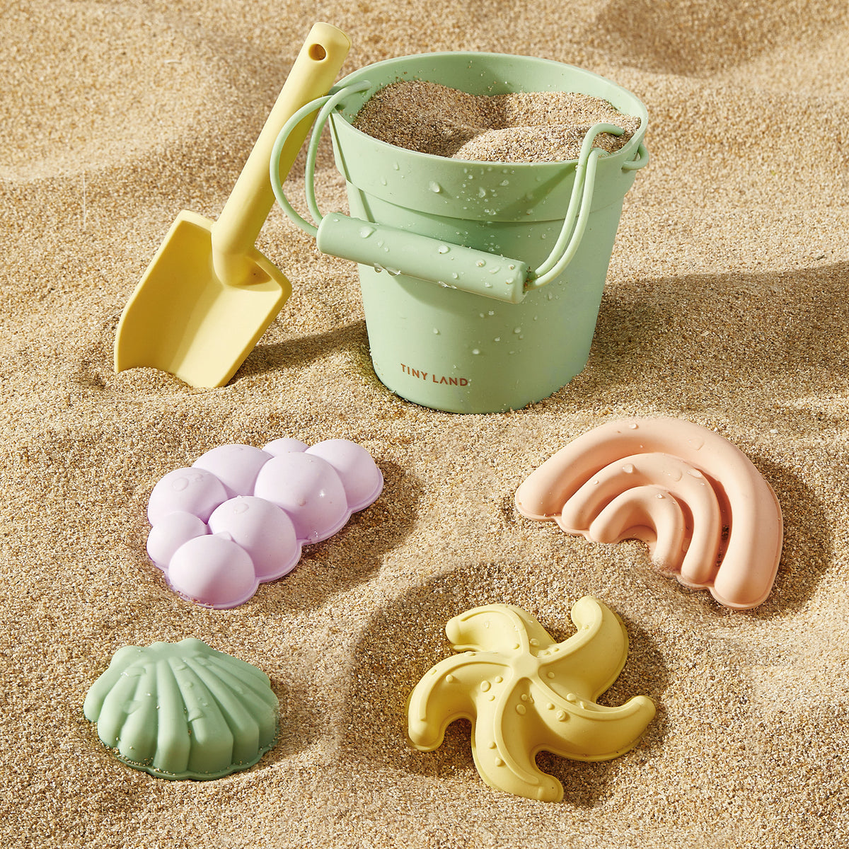 The Tiny Land® Sand-Buddies Sand Toy Set by Tiny Land, featuring a green bucket, yellow plastic shovel, and four pastel sand molds, sits on the sand—perfect for sensory and creative play at the beach.