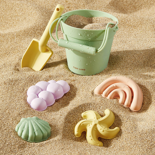 The Tiny Land® Sand-Buddies Sand Toy Set by Tiny Land, featuring a green bucket, yellow plastic shovel, and four pastel sand molds, sits on the sand—perfect for sensory and creative play at the beach.