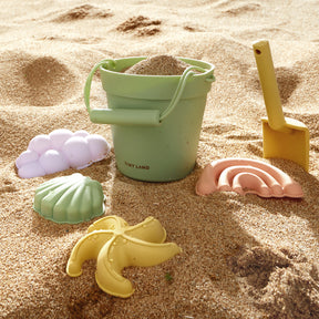 The Tiny Land® Sand-Buddies Sand Toy Set includes a green bucket with sand, a yellow shovel, and four pastel silicone molds—banana, seashell, shell, and rainbow—for endless toddler fun at the beach.