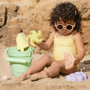 A young child in a yellow-striped swimsuit and sunglasses sits on the sandy beach, playing with the Tiny Land® Baby Beach Tent & Sand Toy Set by Tiny Land and a green bucket, with rocks in the background.|Pink