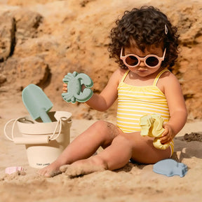 A toddler wearing a pastel yellow striped swimsuit and pink sunglasses plays on the sand with the Tiny Land® One-Stop Beach Toy & Tent Set, featuring a beige bucket and green shovel, with rocks in the background.|Pastel Yellow Stripes
