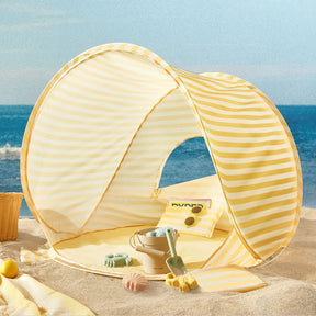 The Tiny Land® One-Stop Beach Toy & Tent Set in pastel yellow stripes sits on the sand, complete with a pillow and sand play set—bucket, shovel, and molds—by the sea under a blue sky.|Pastel Yellow Stripes
