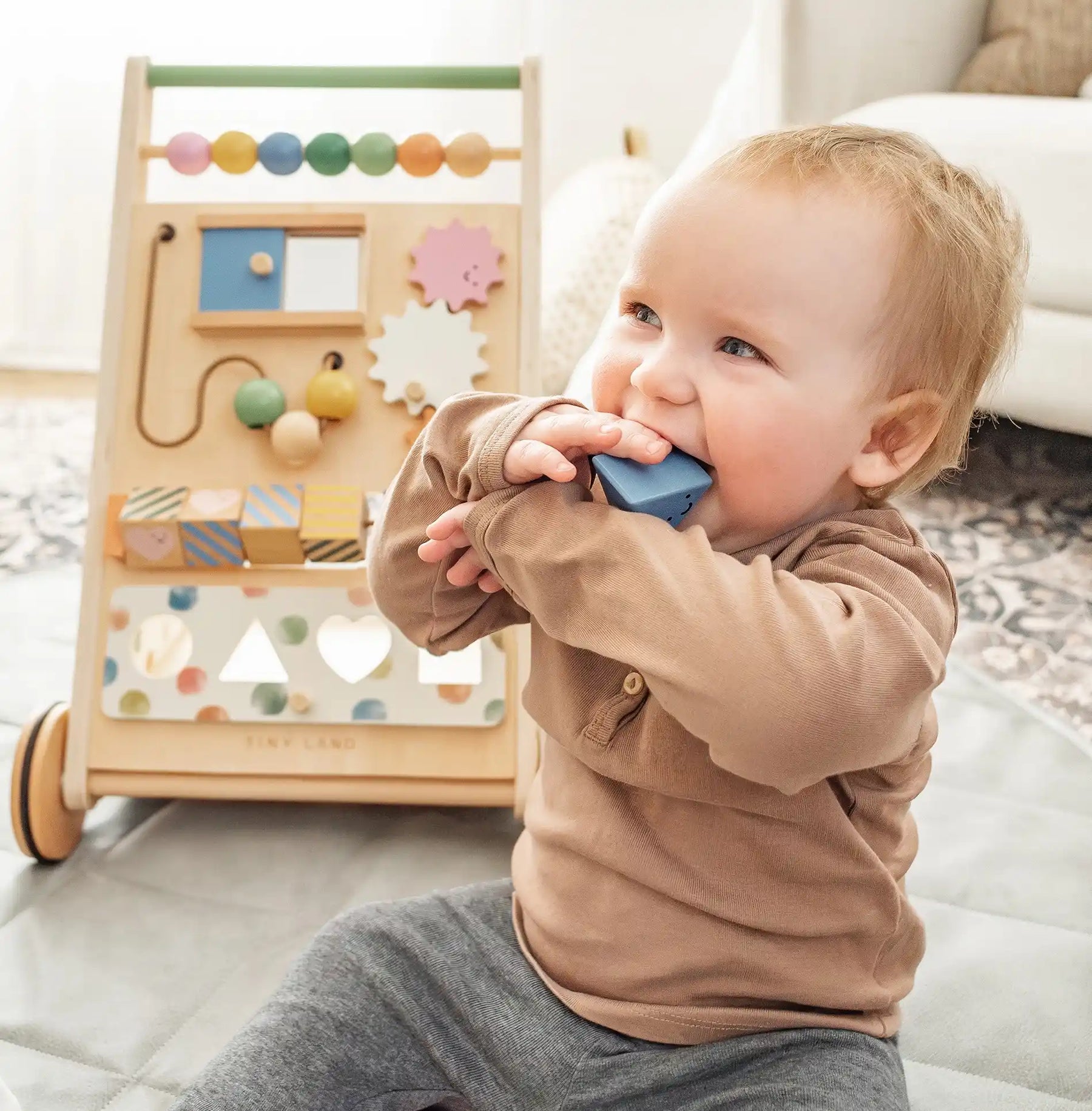A baby with light hair and a brown shirt sits chewing on a blue wooden block. Behind them is the Tiny Land® Versatile Natural Wooden Baby Walker by Tiny Land, featuring colorful beads and gears, indoors on a patterned rug|Morandi