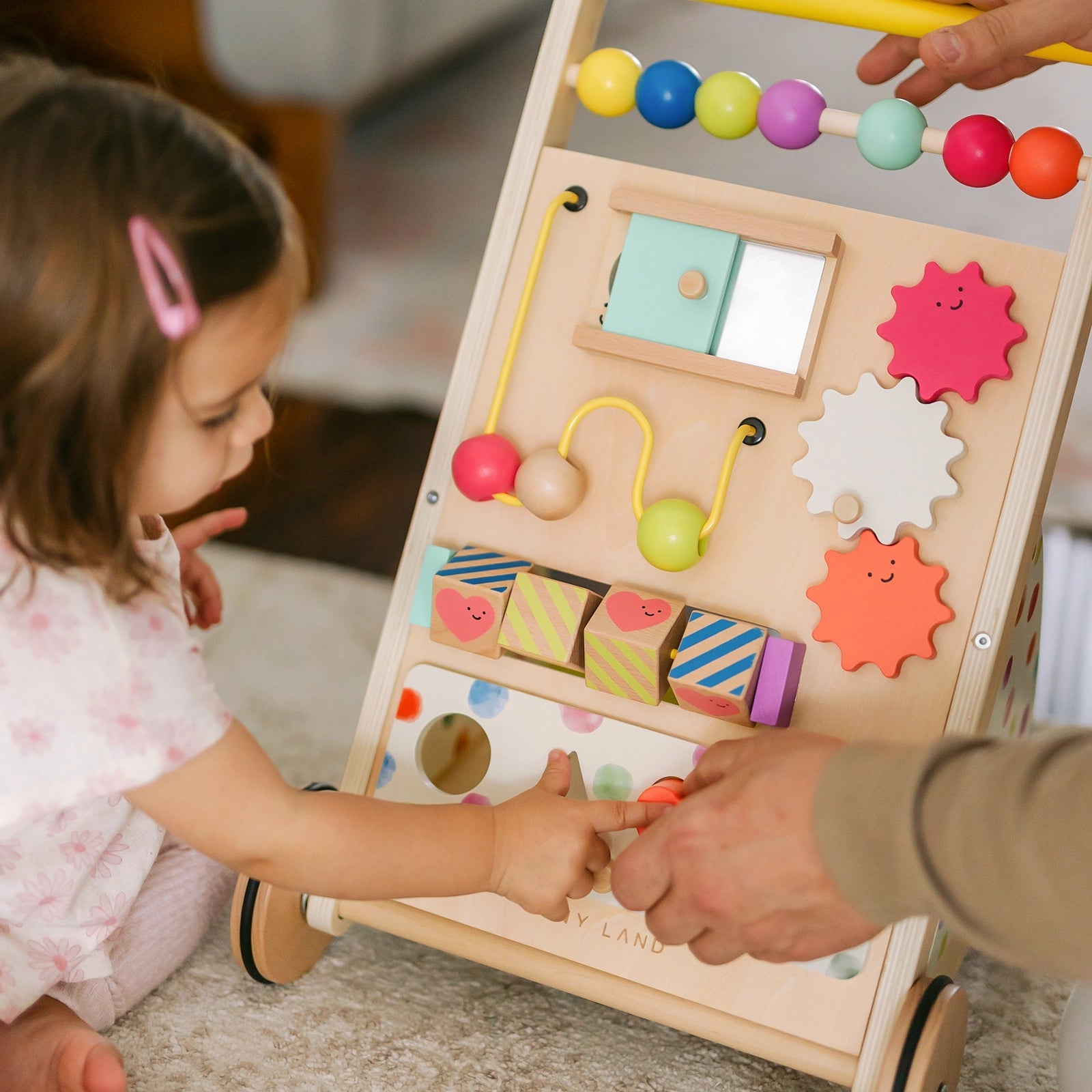 A young child explores the Tiny Land Rainbow Baby Walker & Activity Cube, featuring colorful beads, gears, and blocks. An adult’s hand guides the playset to help develop the child's fine motor skills|Rainbow