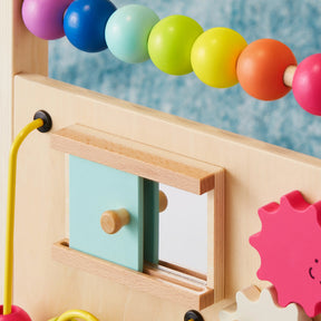 A close-up of the Tiny Land Rainbow Baby Walker & Activity Cube Developmental Playset, showing colorful beads, pink gears, and a blue sliding mirror door on a wooden board, set against a blue textured background for fine motor skills development|Rainbow