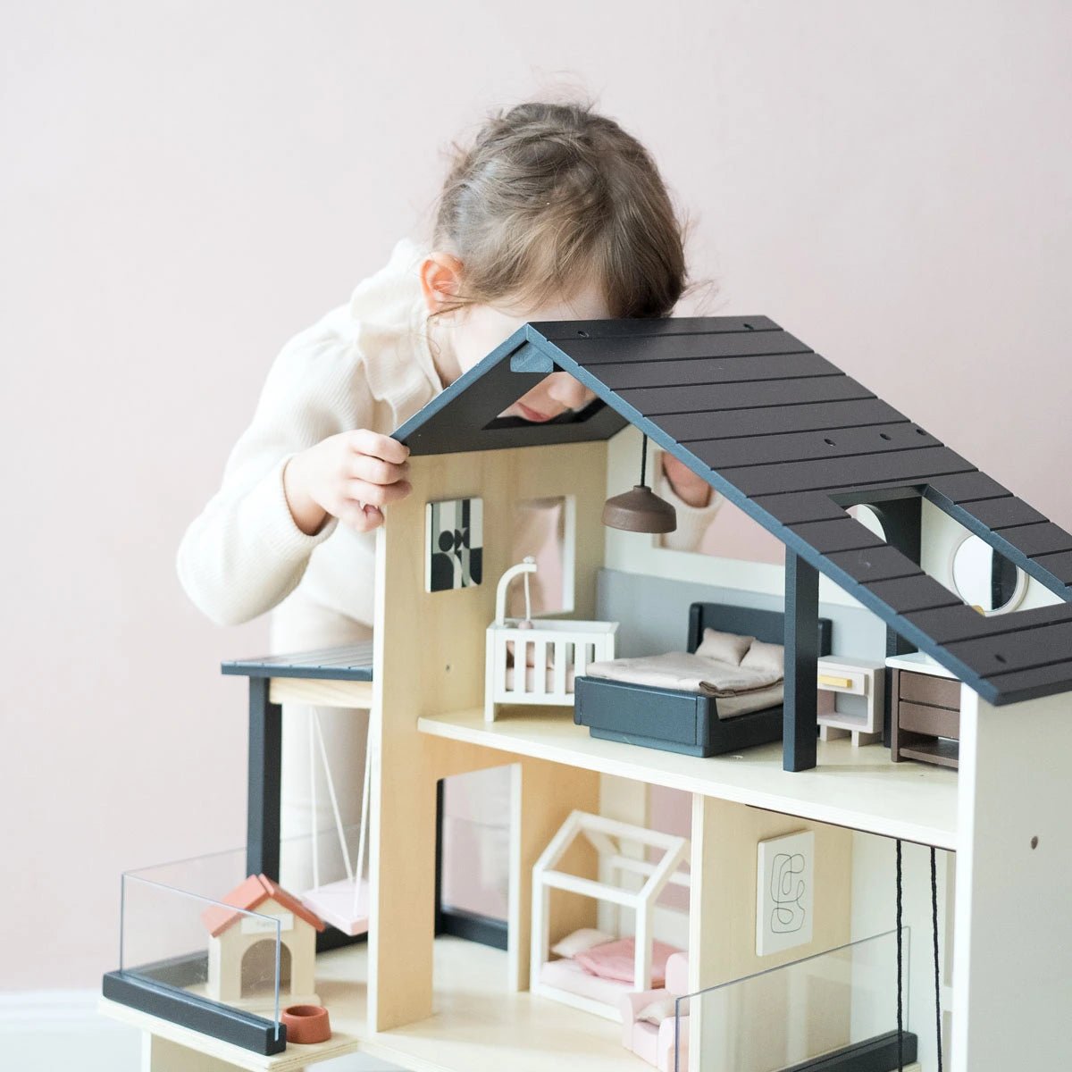 A young child with light brown hair, in a light sweater, leans over to look inside the Tiny Land® Sweetwood Edge Dollhouse by Tiny Land, featuring miniature furniture and family models against a pale pink background.