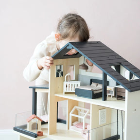 A young child with light brown hair, in a light sweater, leans over to look inside the Tiny Land® Sweetwood Edge Dollhouse by Tiny Land, featuring miniature furniture and family models against a pale pink background.