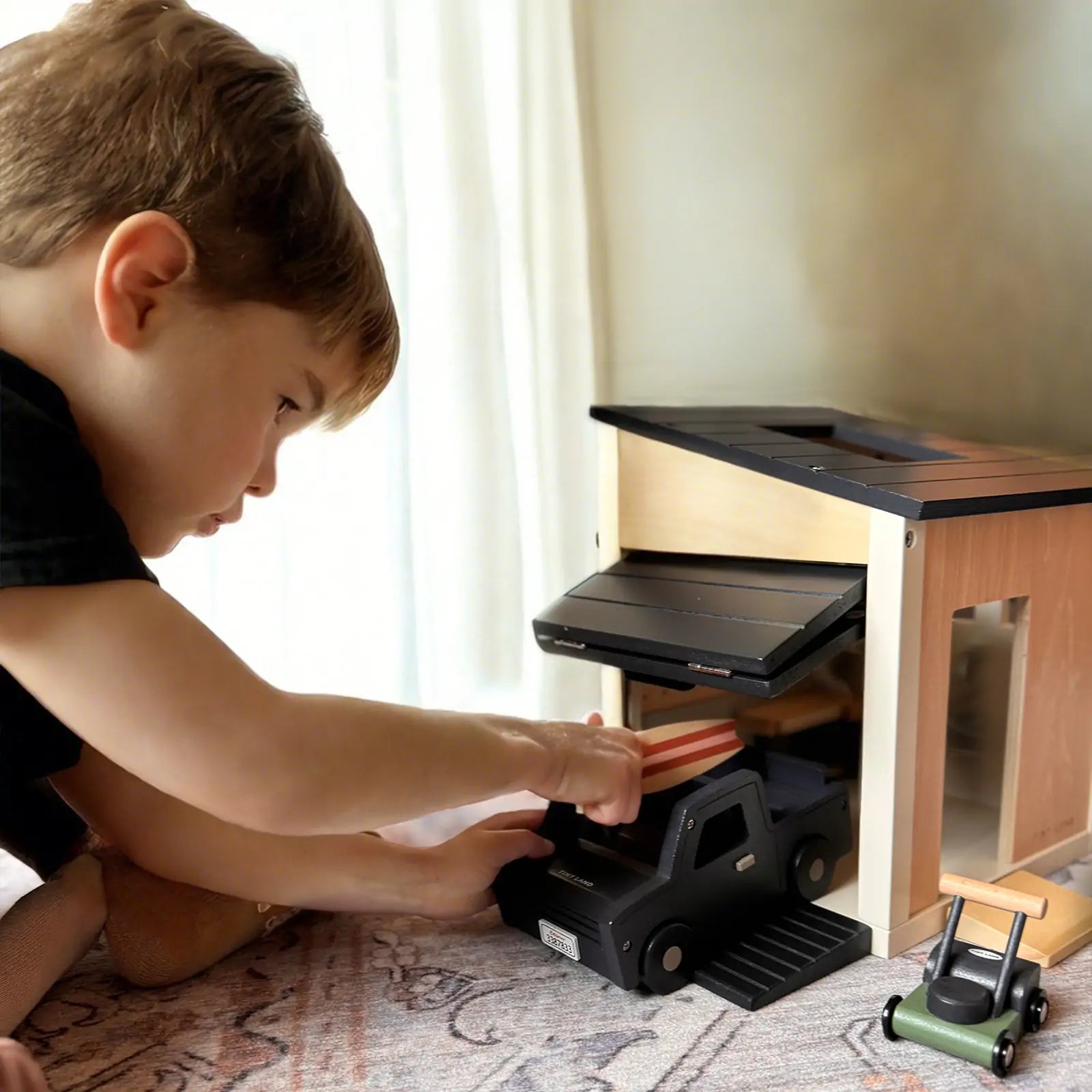 A young boy plays on the floor with the Tiny Land® Wooden Dollhouse - Garage, parking a black toy truck inside while a green toy lawnmower sits nearby. Sunlight streams through a curtain in the background.